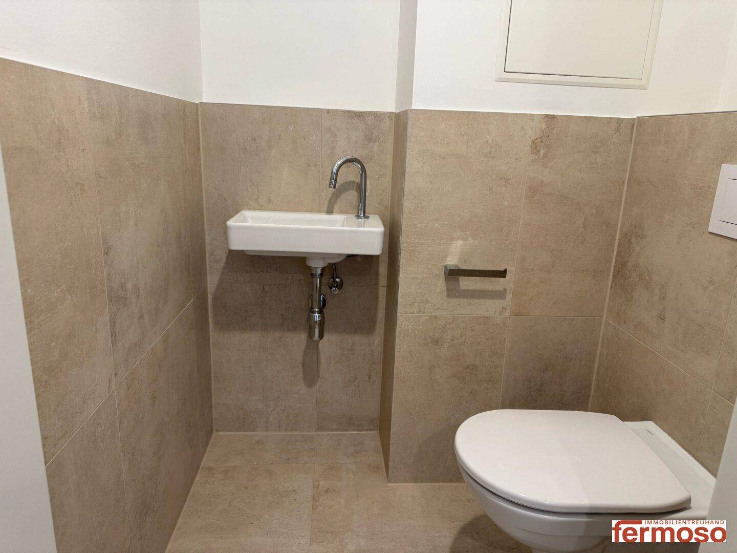 Separate Toilette mit beigefarbenen Fliesen und kleinem Waschbecken.