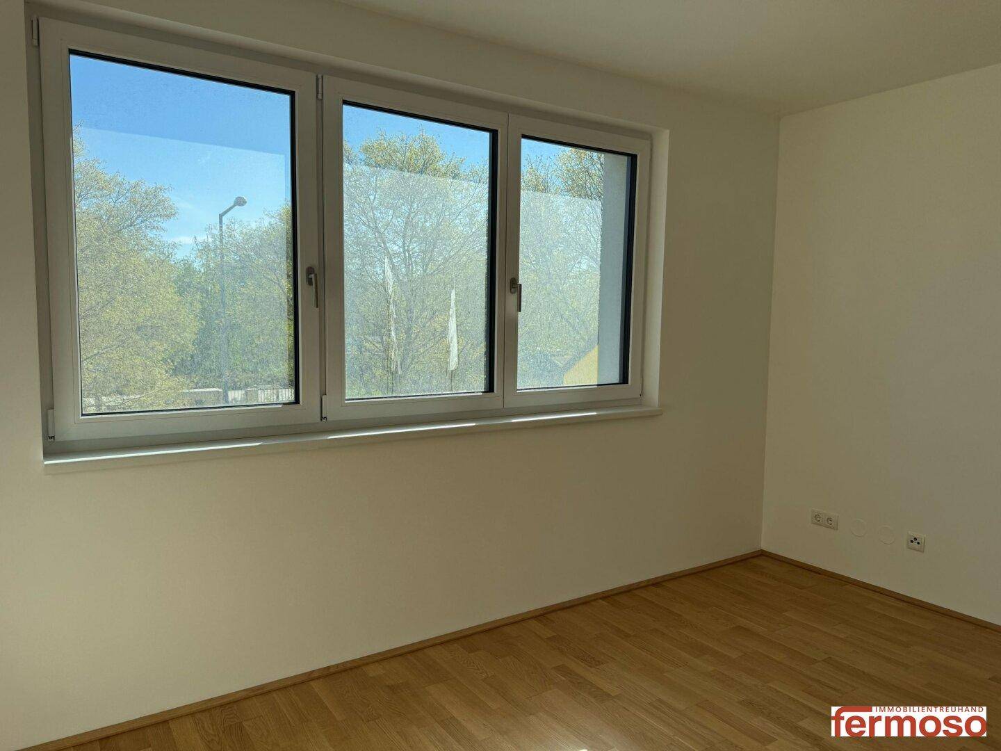 Helles Schlafzimmer mit Parkettboden und Blick ins Grüne durch große Fenster.