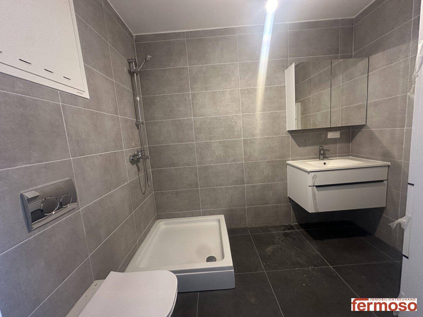 Modernes Badezimmer mit grauen Fliesen, Dusche, WC und Waschtisch mit Spiegelschrank.