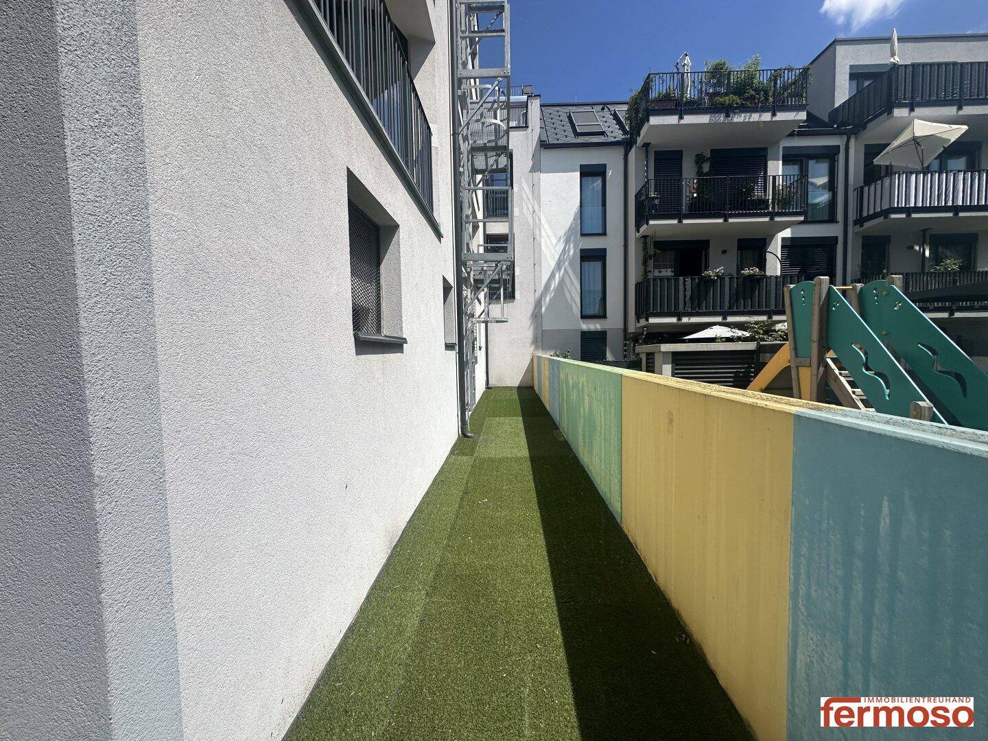 Schmaler Grünstreifen neben dem Gebäude mit Blick auf einen Spielplatz.