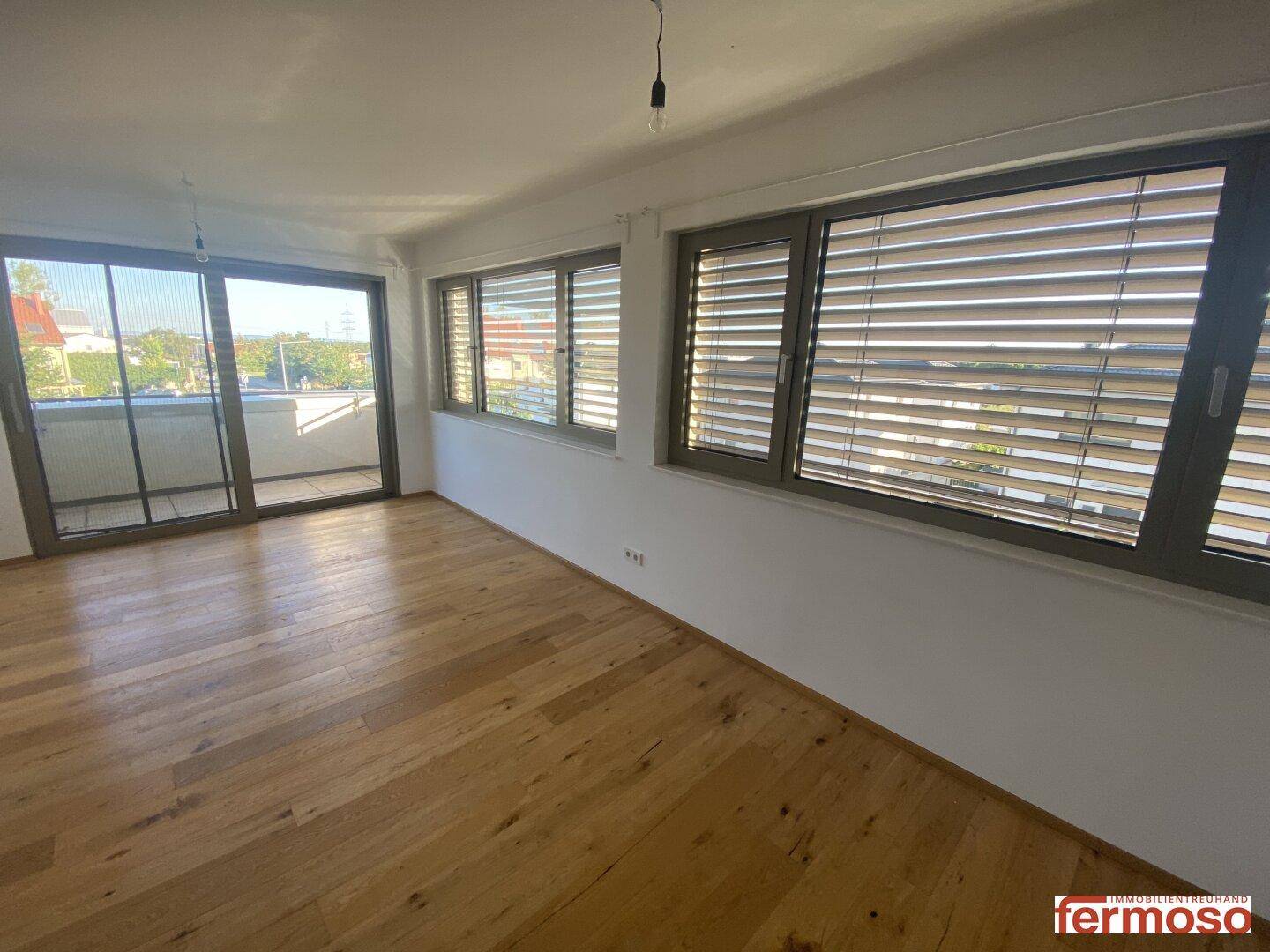 Leeres Wohnzimmer mit Holzboden und großen Fenstern, die viel Licht hereinlassen.