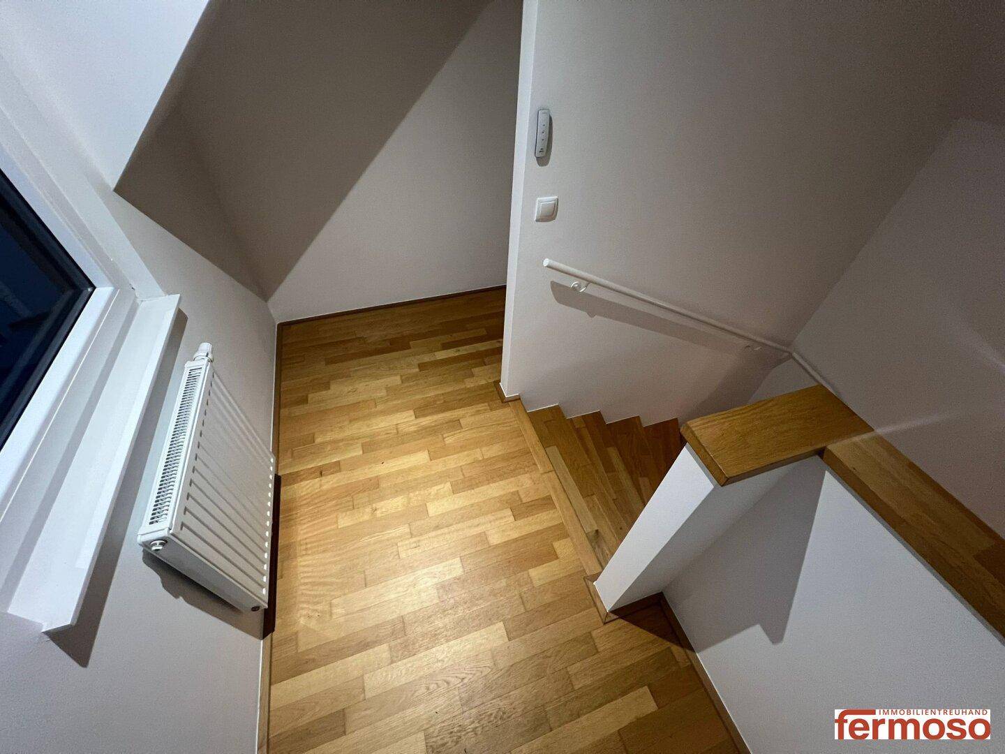 Treppenbereich mit Holzstufen, Parkettboden und Heizkörper unter einem Fenster.