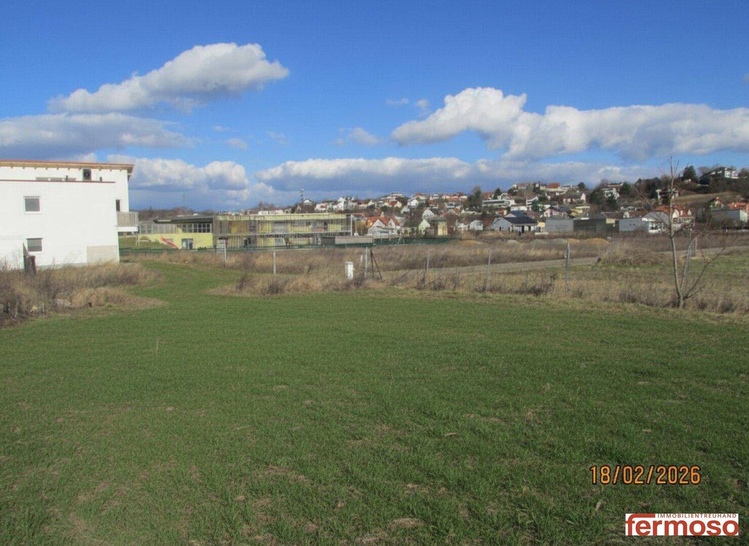 Grüne Wiese mit einem weißen Haus und Blick auf eine Siedlung am Hang unter einem weiten, bewölkten Himmel.