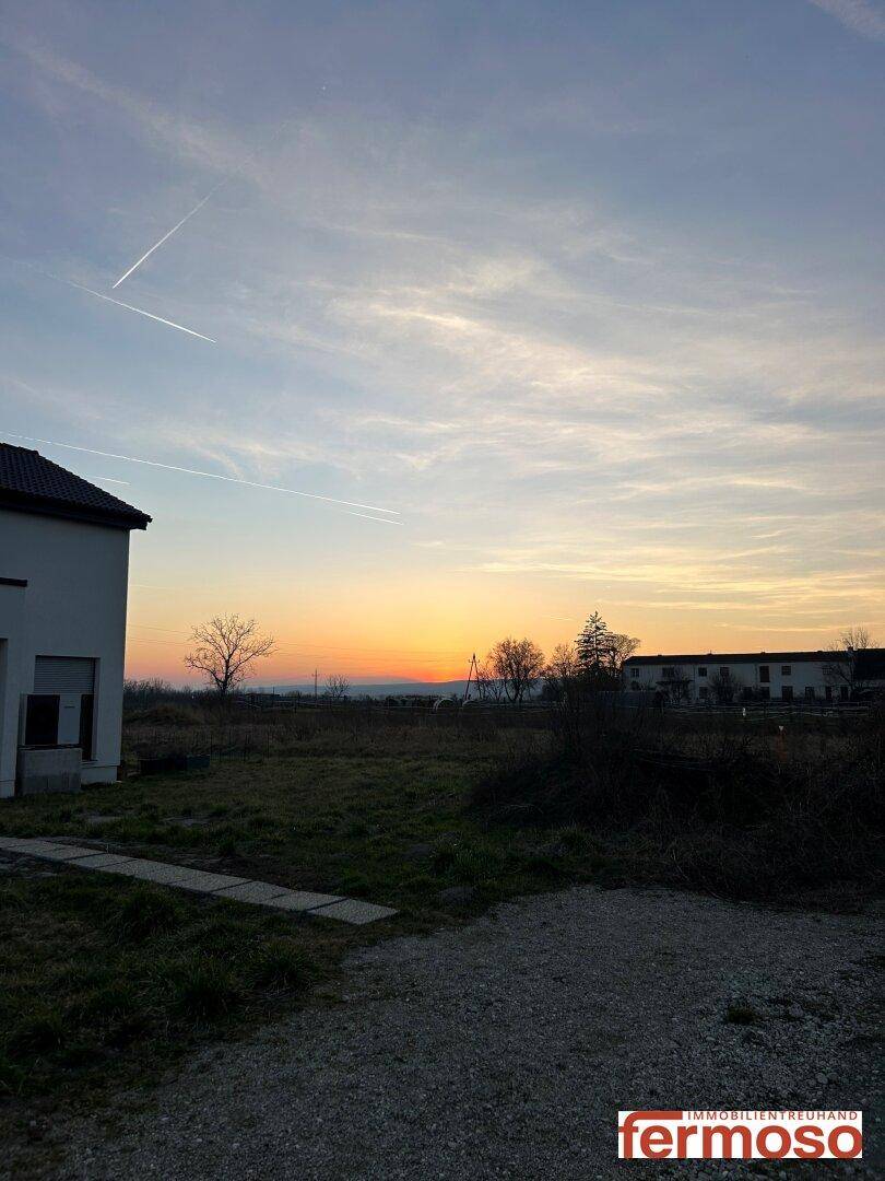 Weißes Haus mit Kiesweg und weitem Feld im Vordergrund, beleuchtet vom warmen Licht des Sonnenuntergangs.