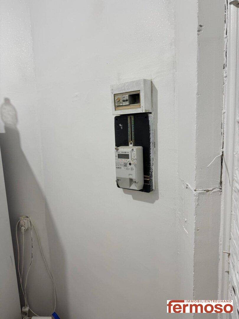 Ein Stromzähler und Sicherungskasten sind an einer weißen Wand in einem Flur montiert, typisch für Versorgungsräume.