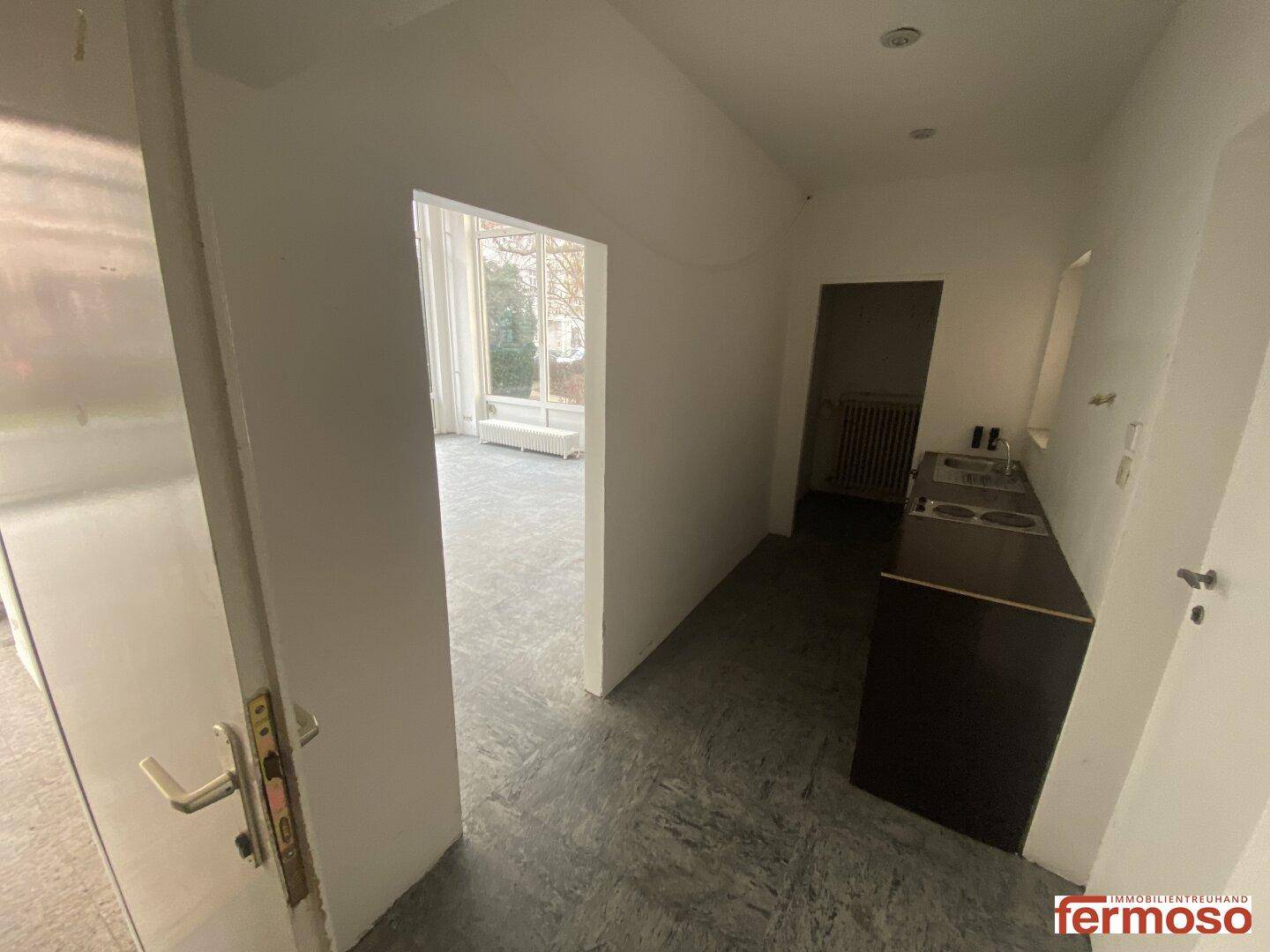 Kleine Küchenzeile mit Spüle und Kochplatten, angrenzend an einen hellen Raum mit großen Fenstern.