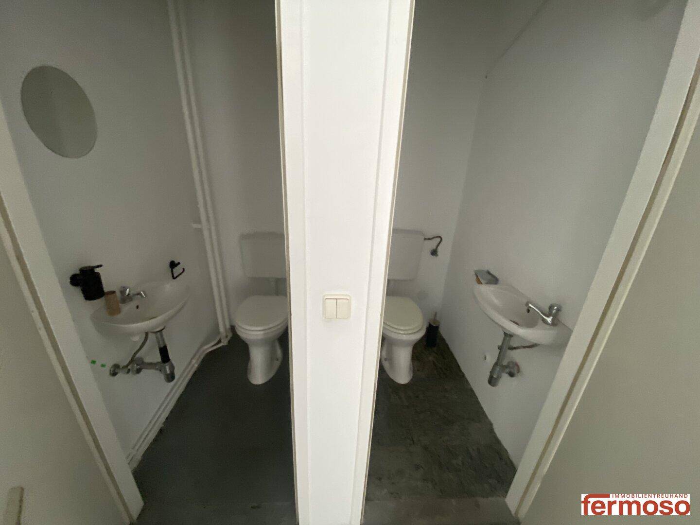 Zwei separate Toilettenräume mit Waschbecken, getrennt durch eine Trennwand.