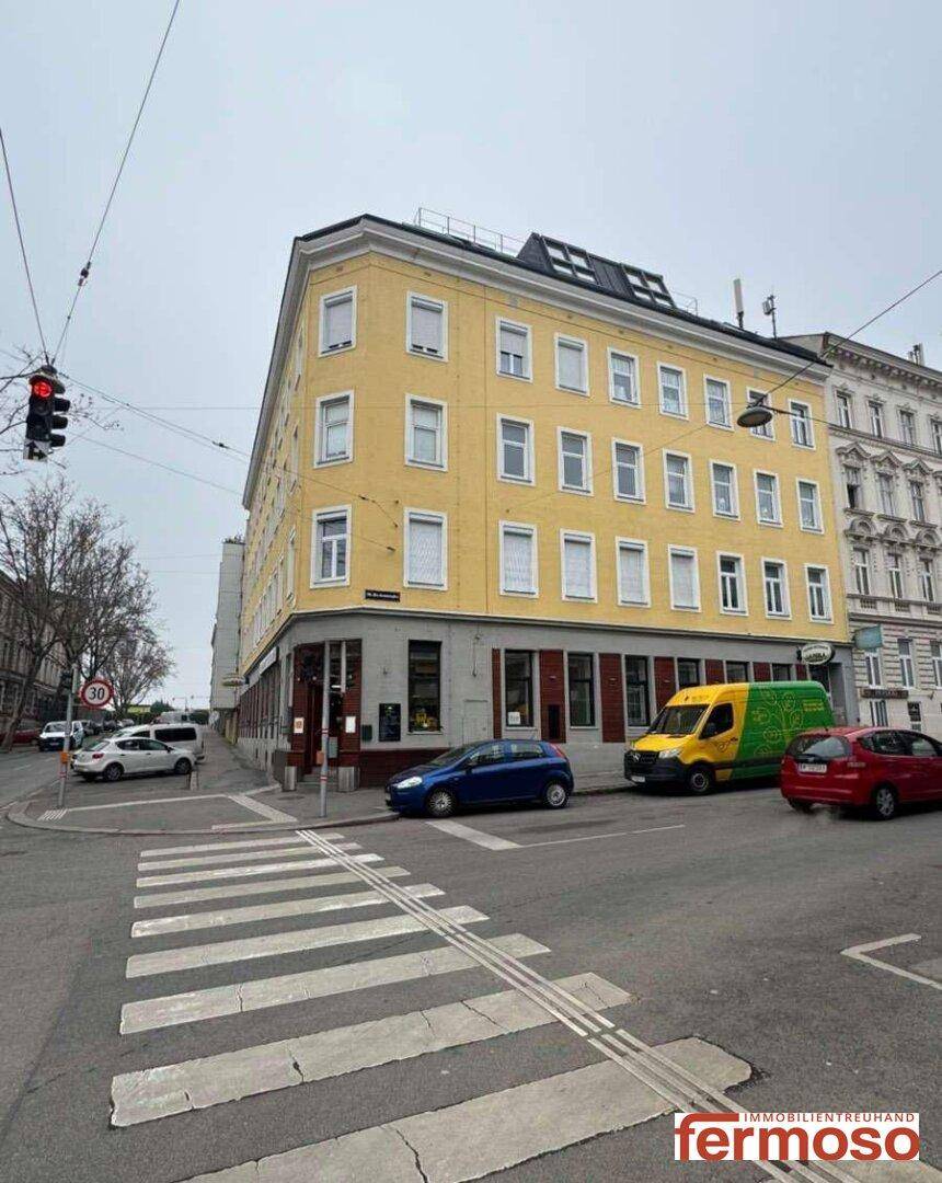 Gelbes Eckgebäude an einer belebten Kreuzung mit mehreren Fenstern und Geschäften im Erdgeschoss.