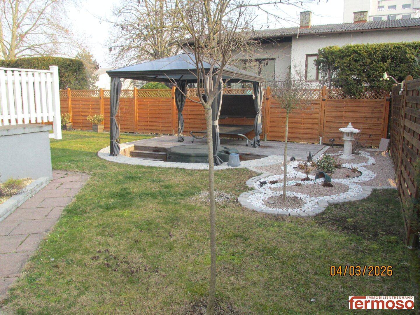 Gepflegter Garten mit Rasenfläche, Pavillon und dekorativen Steinelementen, umgeben von einem Holzzaun.