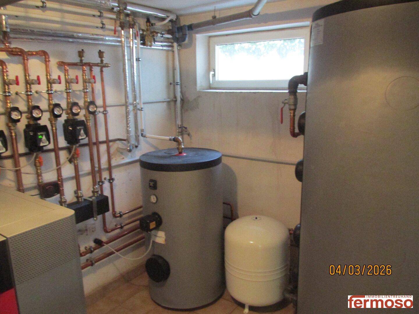 Heizungsanlage mit Warmwasserspeichern und Rohrleitungen in einem Technikraum mit Fenster.