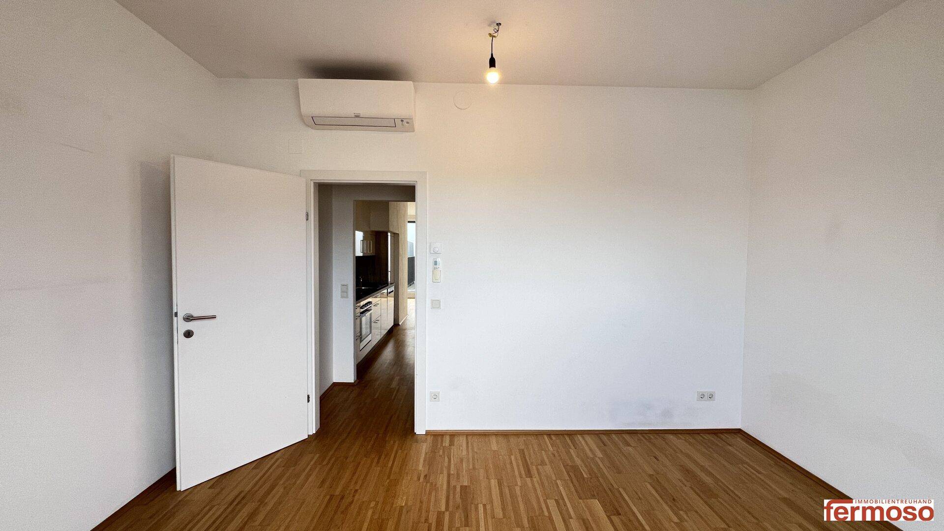Leeres Wohnzimmer mit Holzboden und weiß gestrichenen Wänden, bietet Zugang zur Küche.