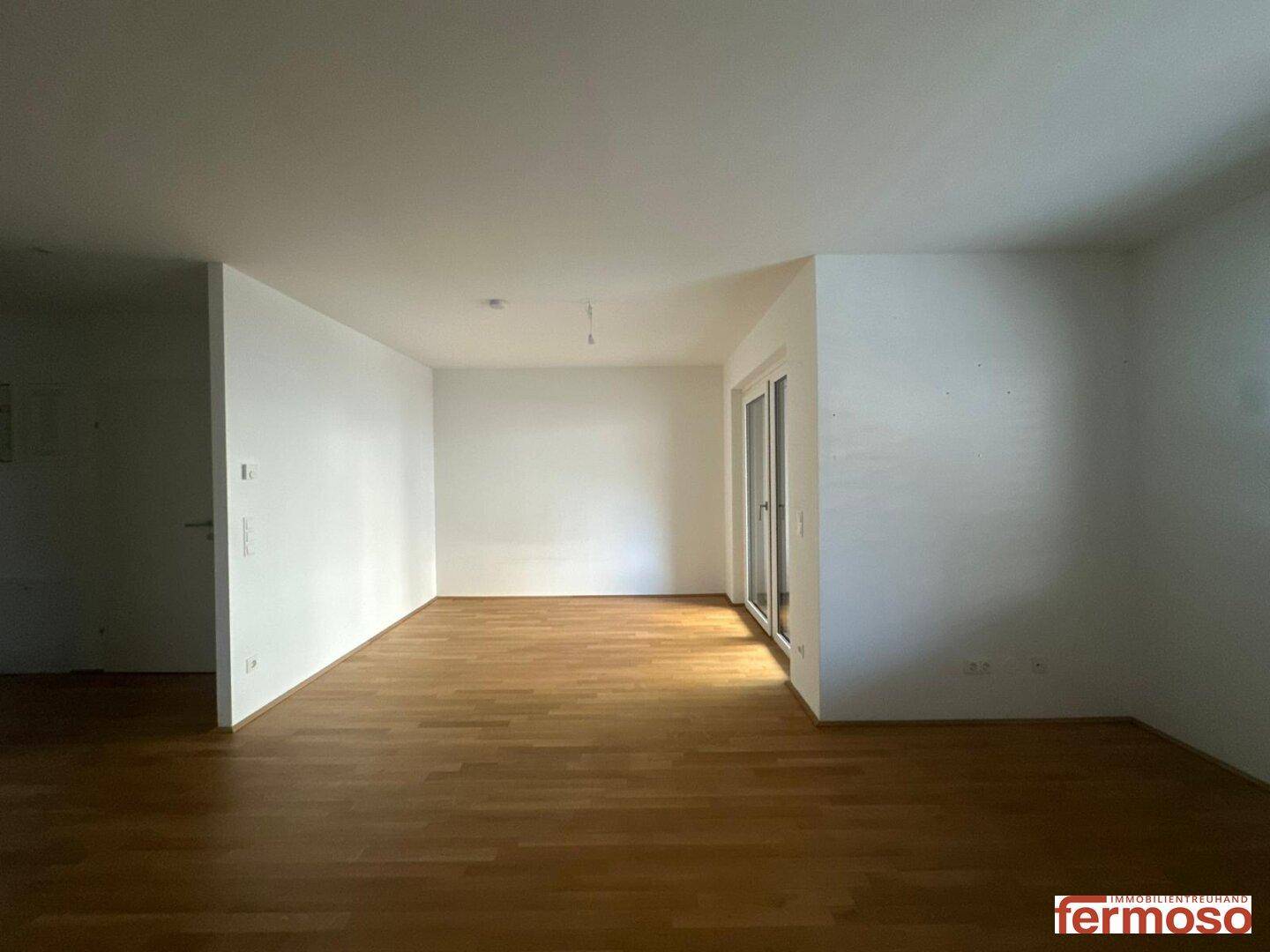 Großer, leerer Wohnbereich mit Parkettboden und viel Tageslicht durch die Balkontür.
