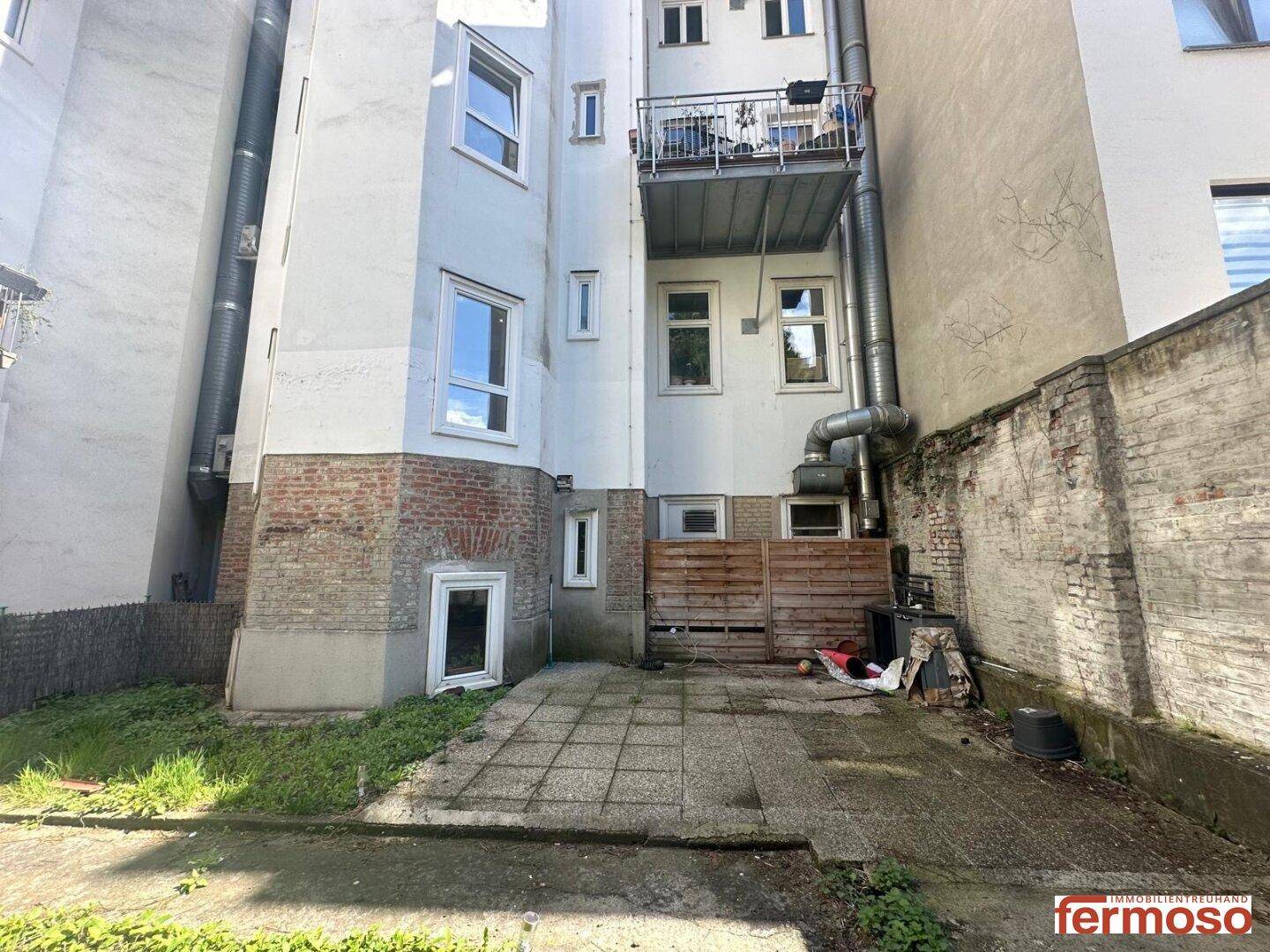 Rückansicht eines Gebäudes mit Balkon, Abluftrohren und einem kleinen gepflasterten Bereich.