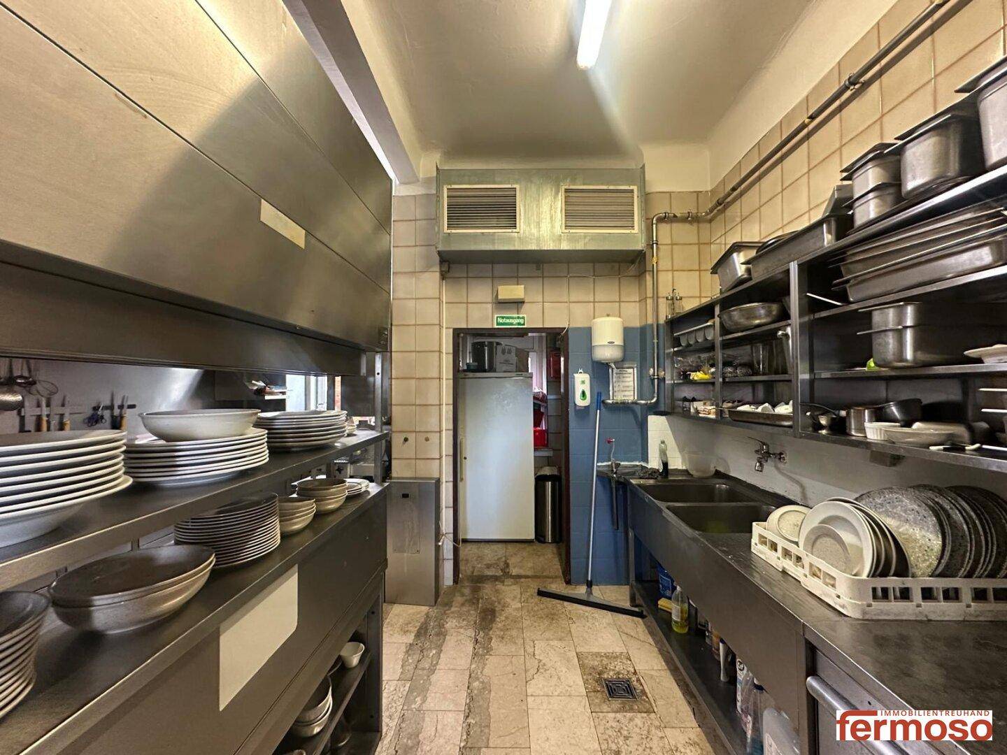 Industrielle Küche mit Edelstahl-Arbeitsflächen, Regalen voller Geschirr und Fliesenboden.