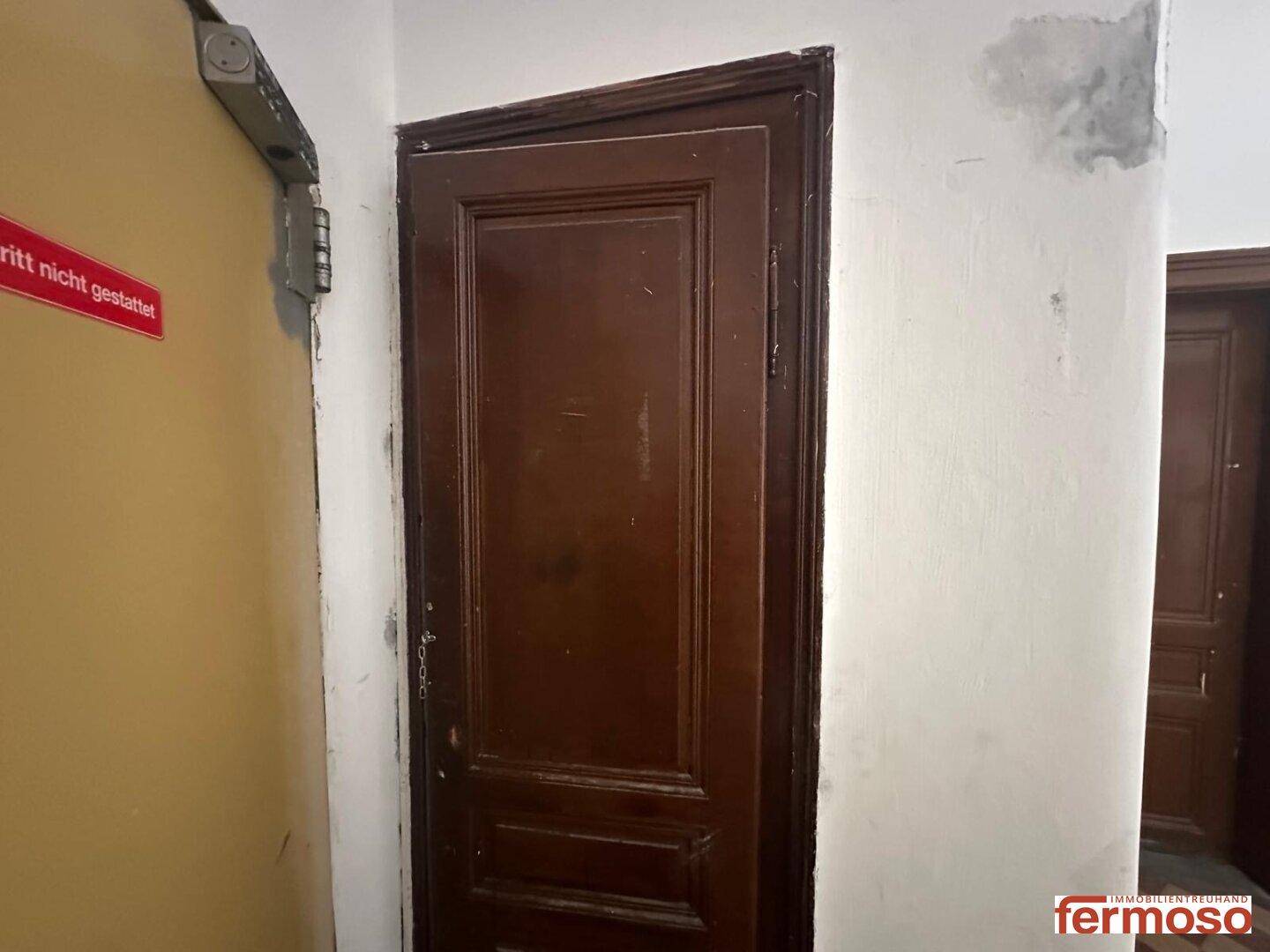Eine alte Holztür in einem Flur mit abgenutzten Wänden und sichtbaren Gebrauchsspuren.
