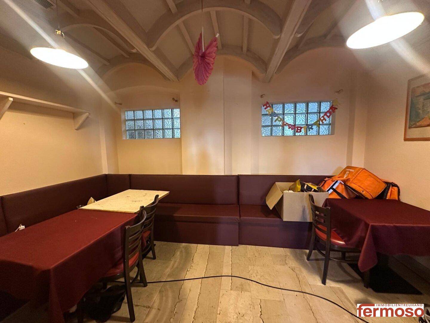 Kellerraum mit Sitzbänken, Tischen und kleinen Glasbausteinfenstern, möglicherweise für Veranstaltungen genutzt.