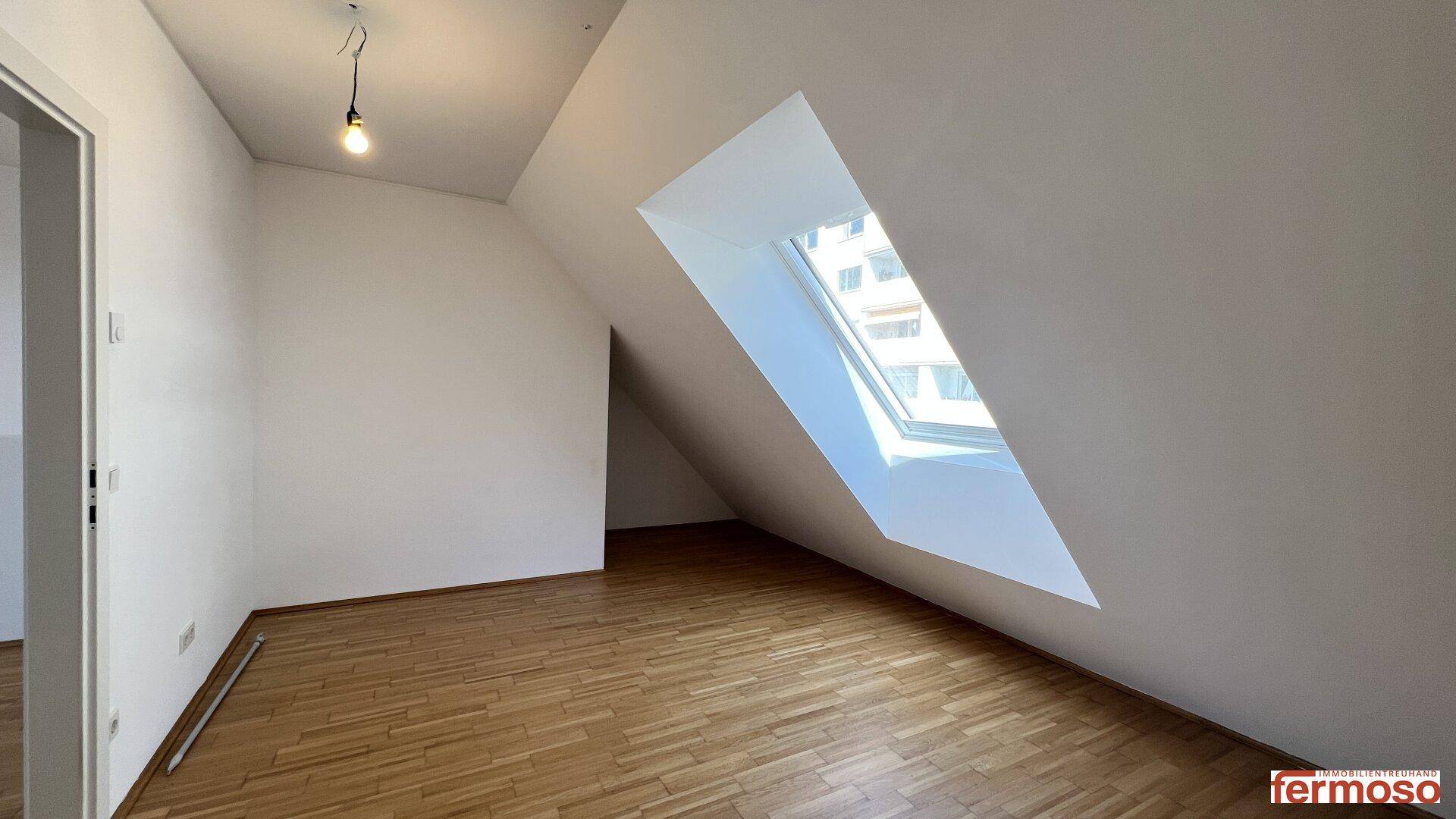 Helles Zimmer im Dachgeschoss mit schrägen Wänden und großem Dachfenster.