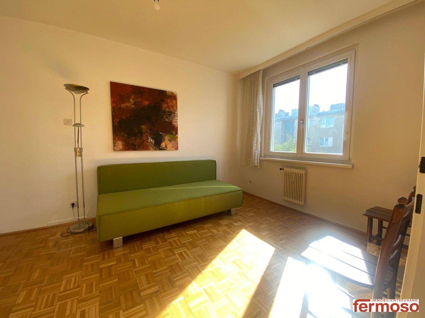 Helles Zimmer mit Parkettboden, grünem Sofa und großem Fenster mit viel Tageslicht.