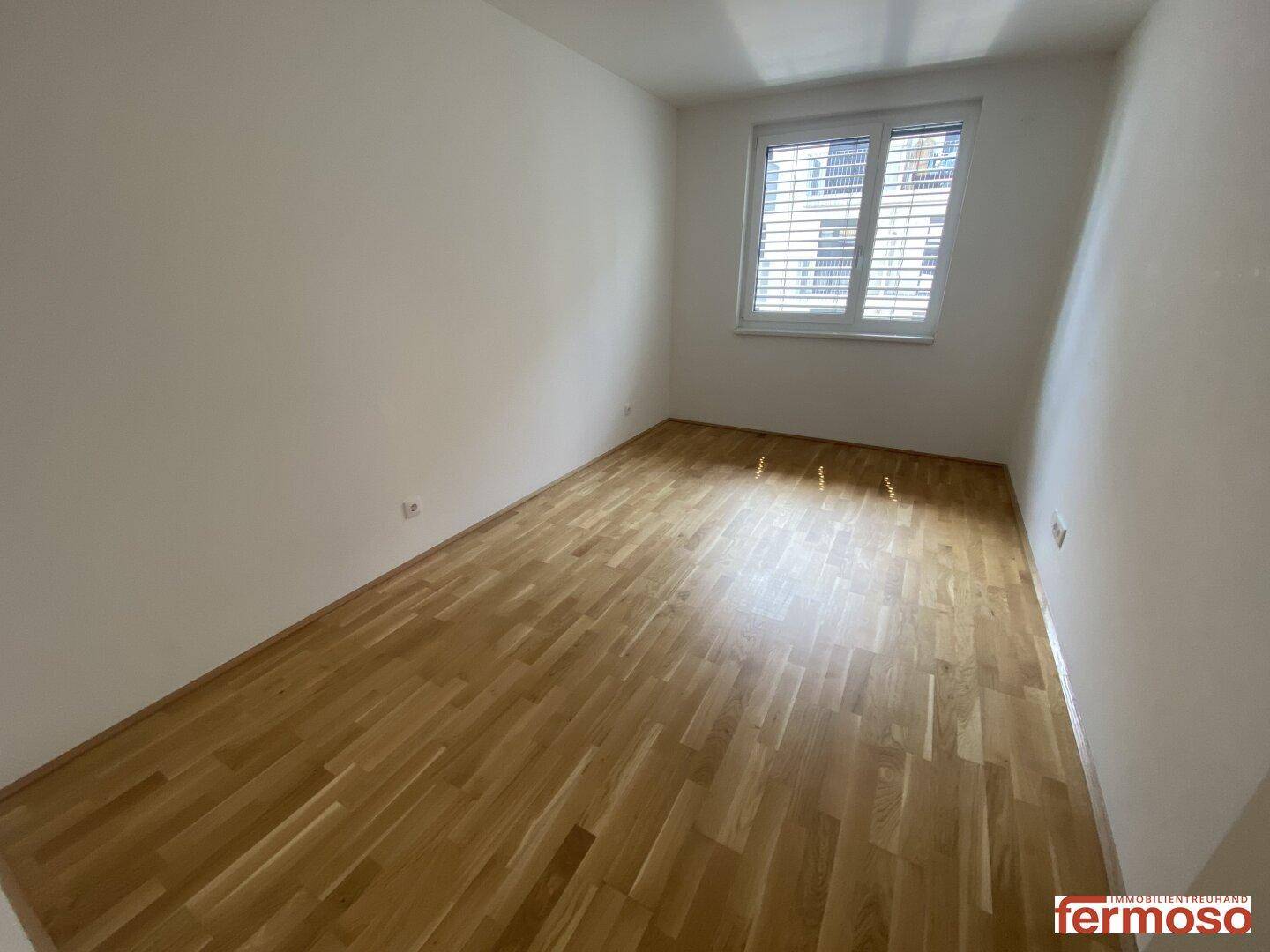 Leeres Schlafzimmer mit Parkettboden und Fenster mit Jalousien, bereit zur individuellen Gestaltung.