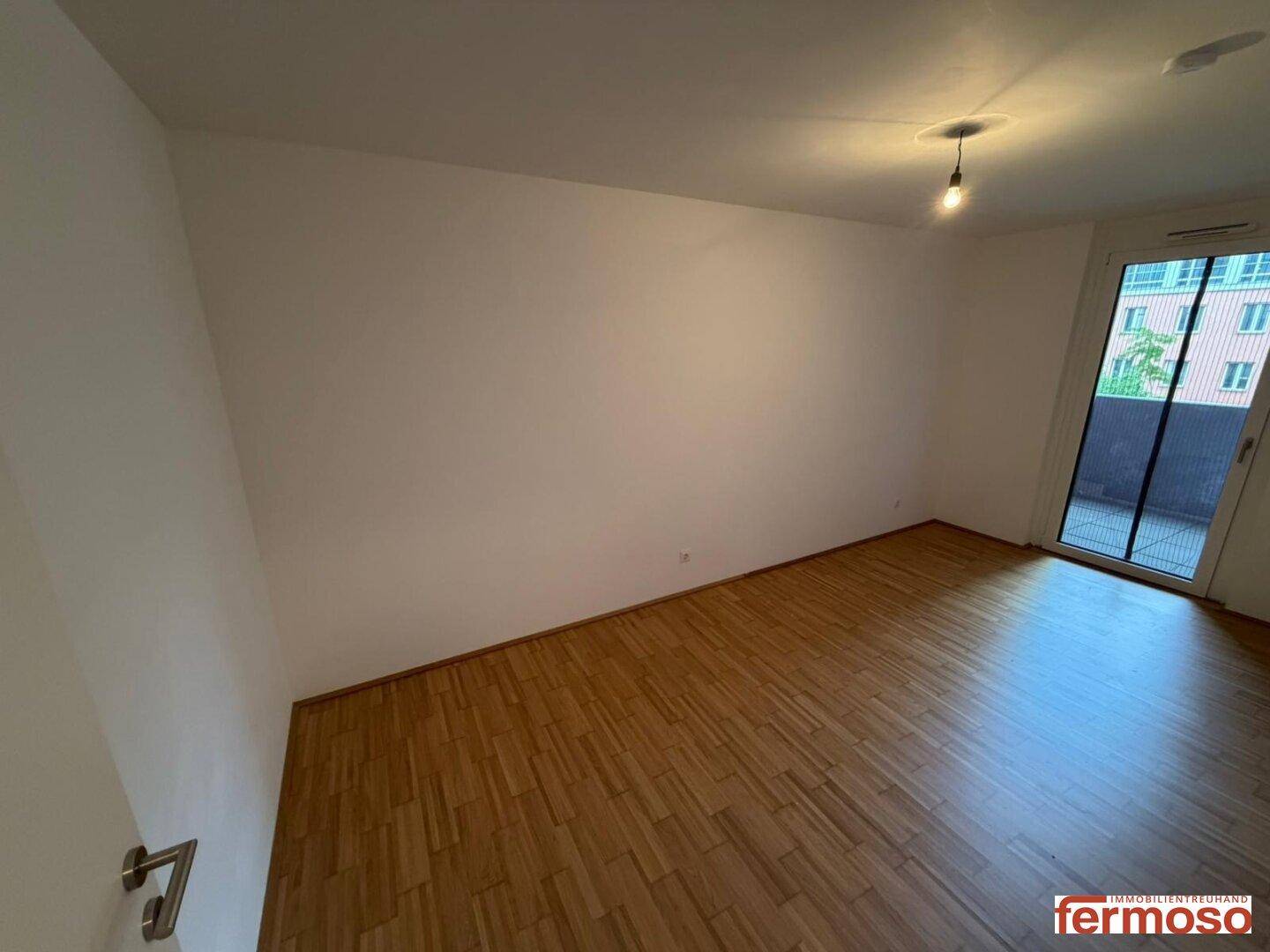 Leeres Zimmer mit Holzboden und direktem Zugang zu einem sonnigen Balkon.