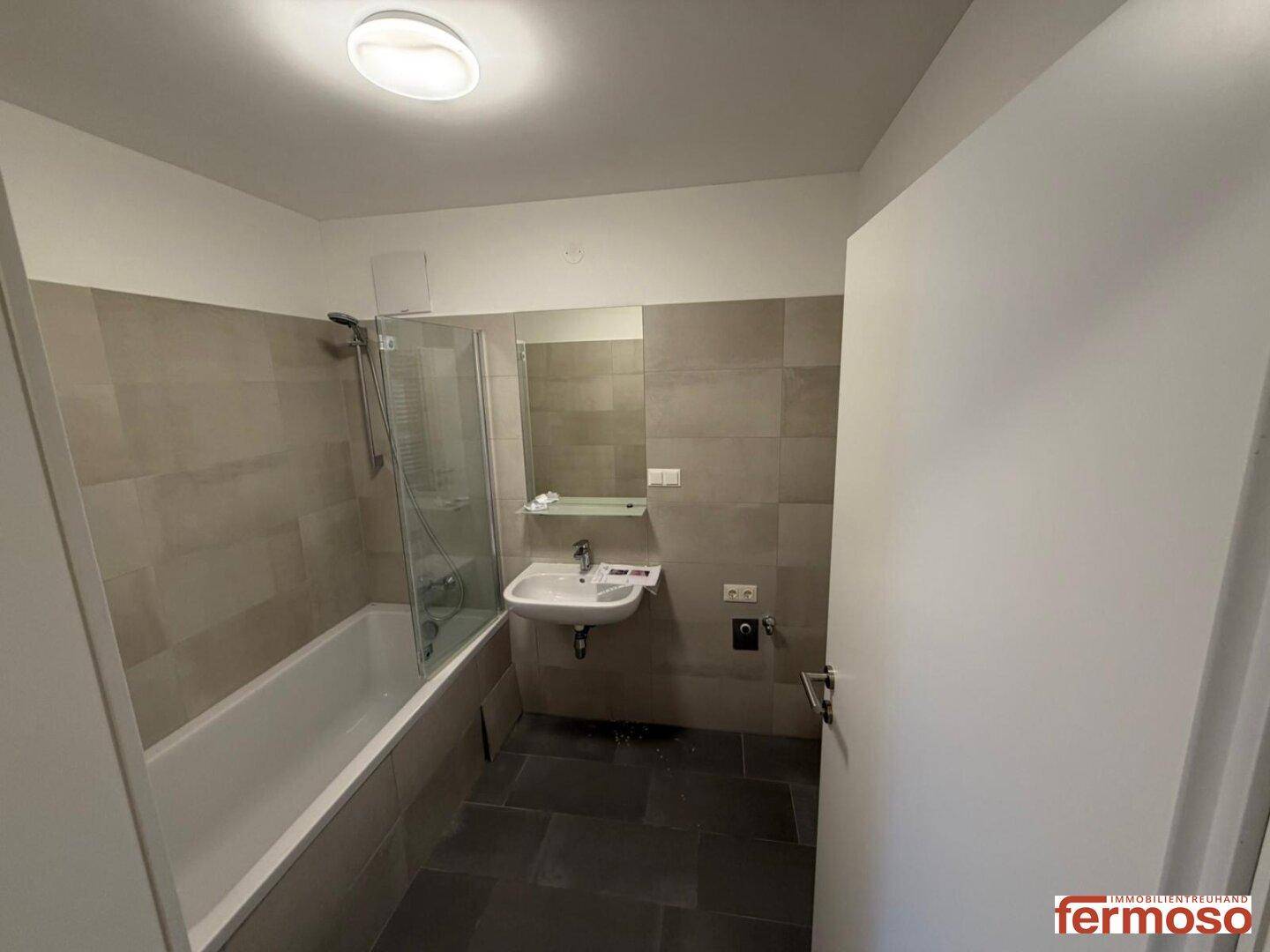 Badezimmer mit Badewanne, Duschabtrennung und modernem Waschbecken, gefliest in hellen Grautönen.