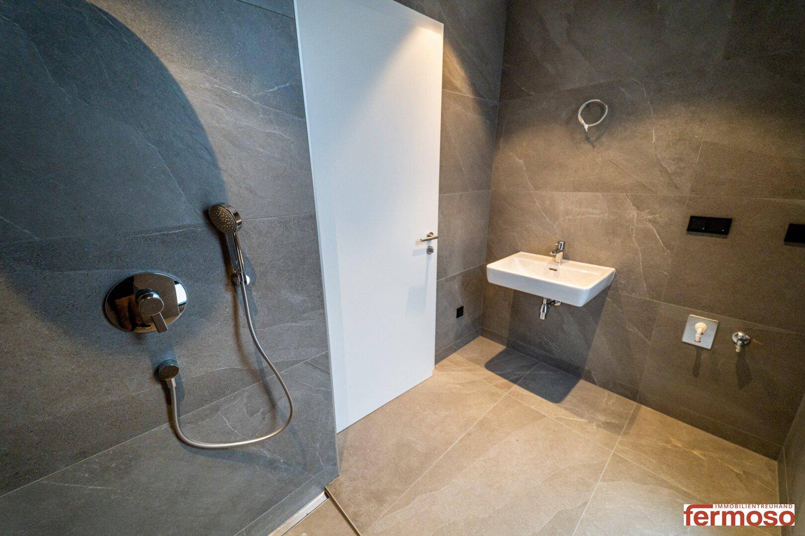 Modernes Badezimmer mit grauen Fliesen, Dusche und wandhängendem Waschbecken.