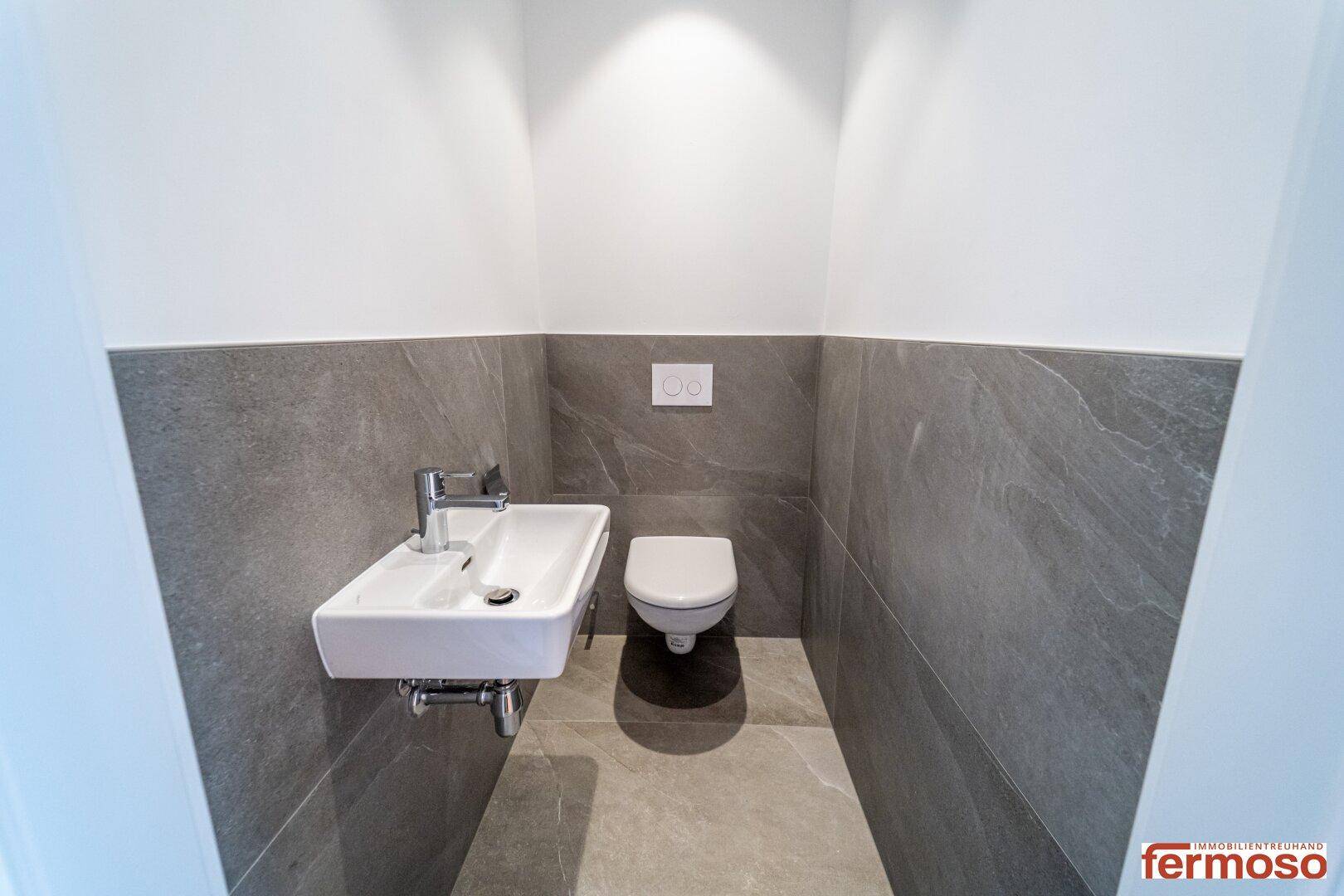 Modernes Gäste-WC mit grauen Fliesen, wandhängendem WC und kleinem Waschbecken.