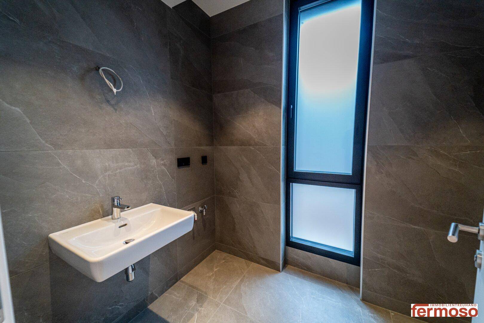 Helles Badezimmer mit grauen Fliesen, Waschbecken und hohem Fenster für Tageslicht.