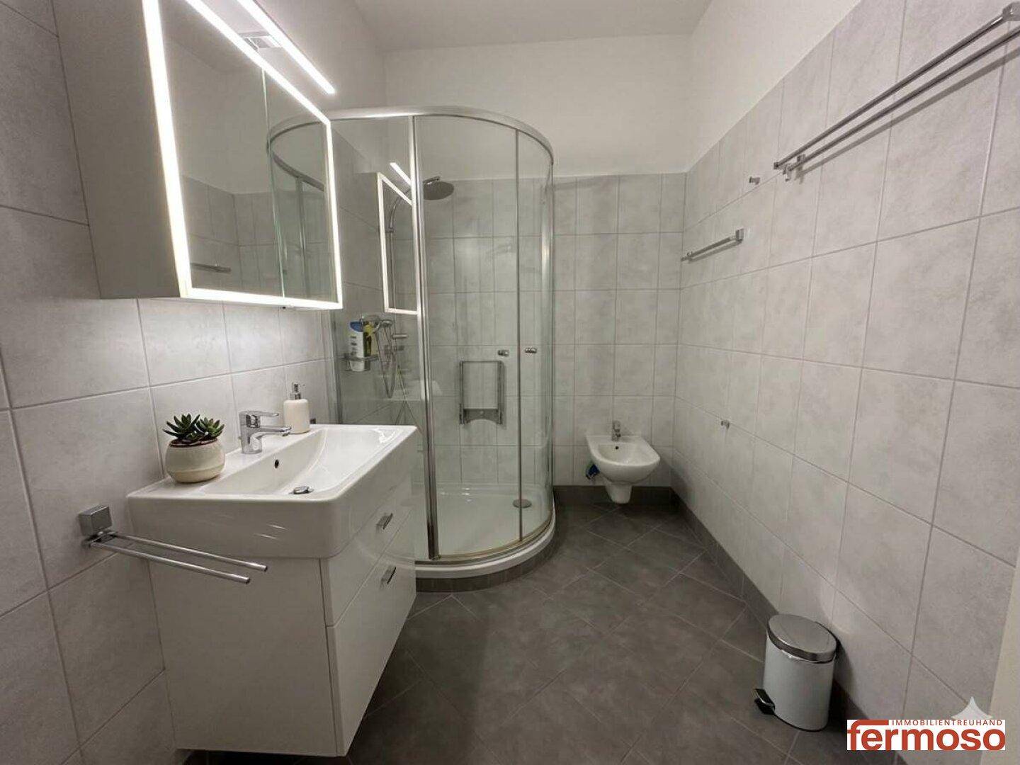 Modernes Badezimmer mit Duschkabine, breitem Waschbecken und Bidet, hell gefliest.