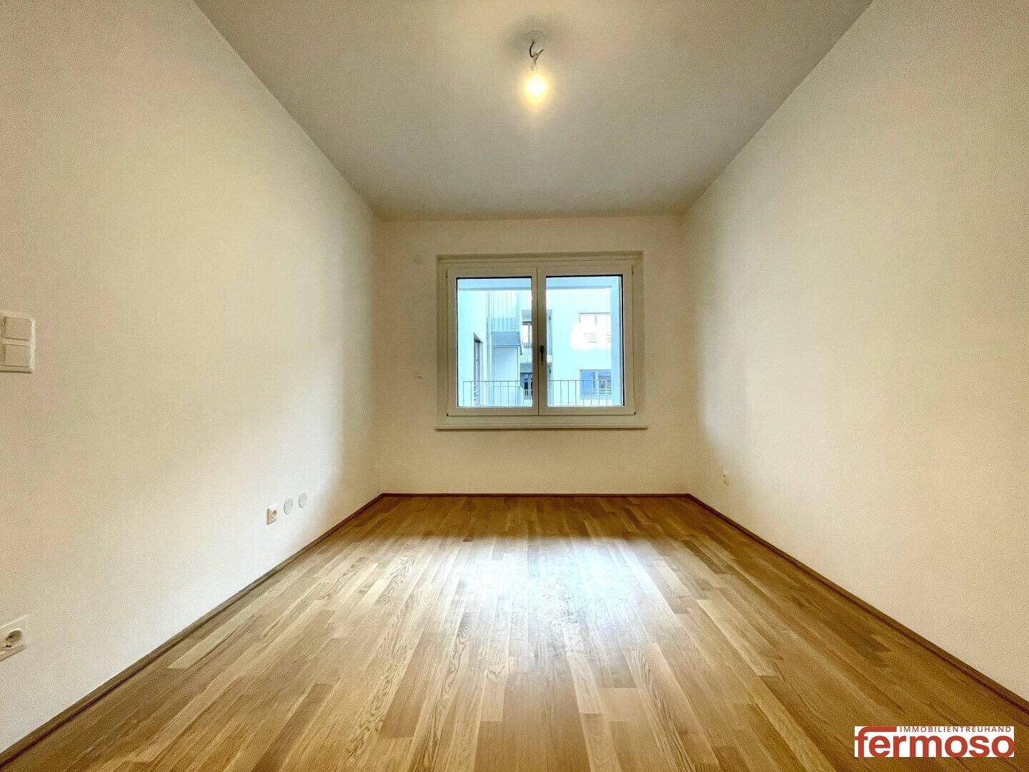 Helles, leeres Zimmer mit Holzboden und Fenster, das einen Blick auf die umliegenden Gebäude bietet.