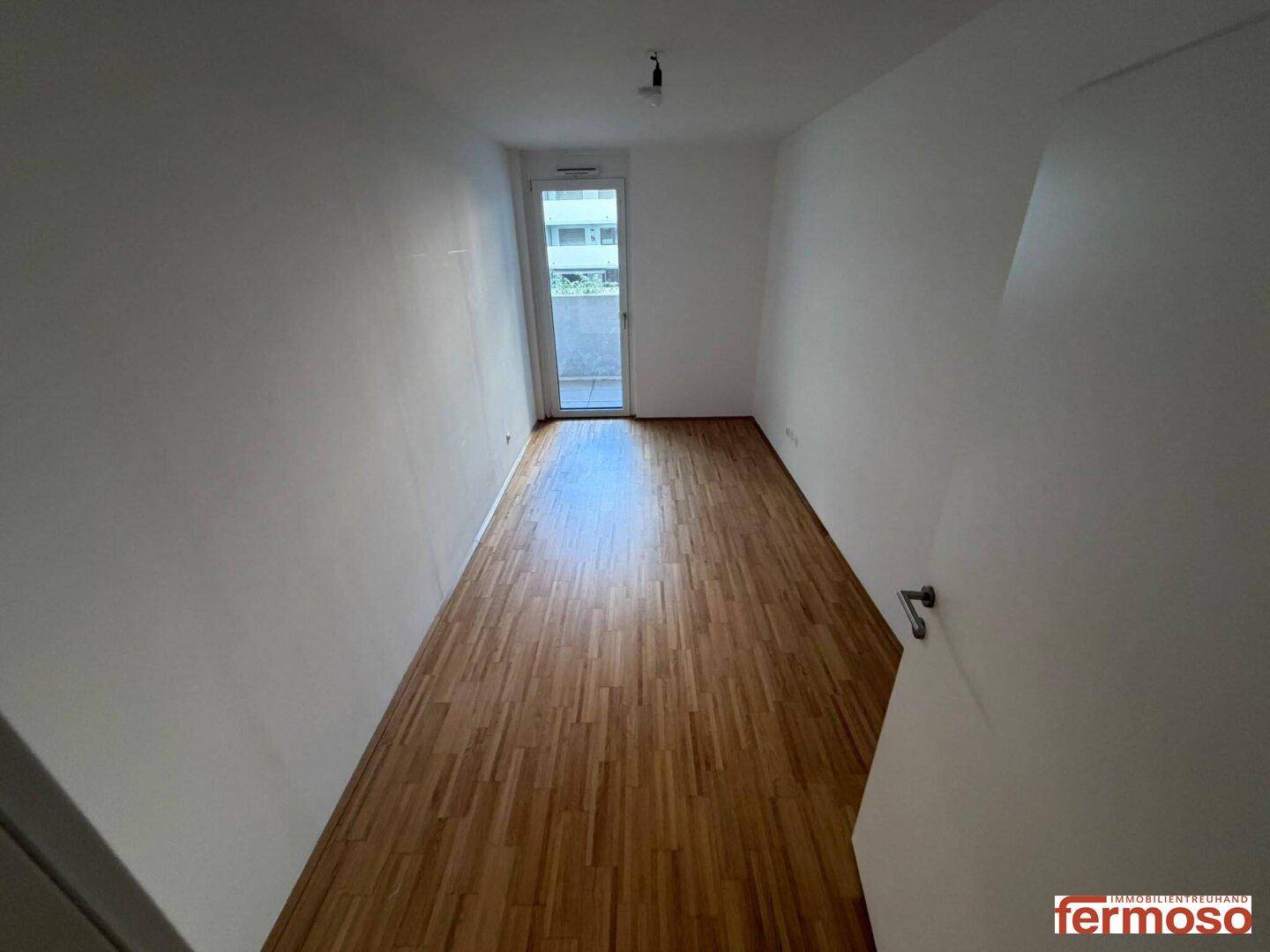 Leeres Schlafzimmer mit hellem Parkettboden und Zugang zu einem Balkon, bietet viel natürliches Licht.