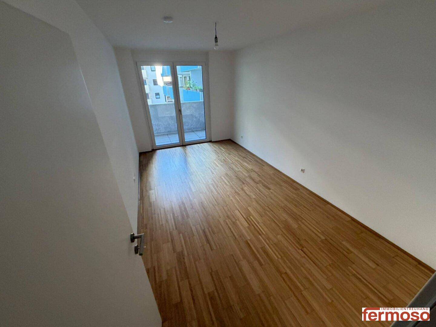 Helles Zimmer mit Holzboden und breiter Balkontür, die viel Tageslicht hereinlässt.