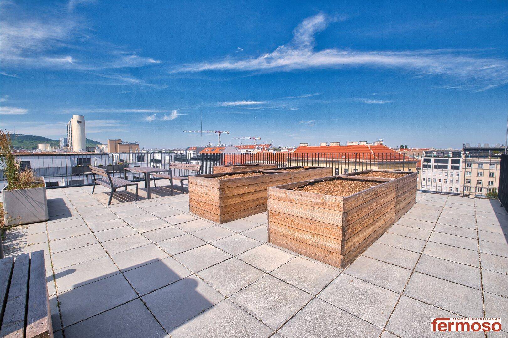 Weitläufige Dachterrasse mit Hochbeeten und Stadtblick unter blauem Himmel.