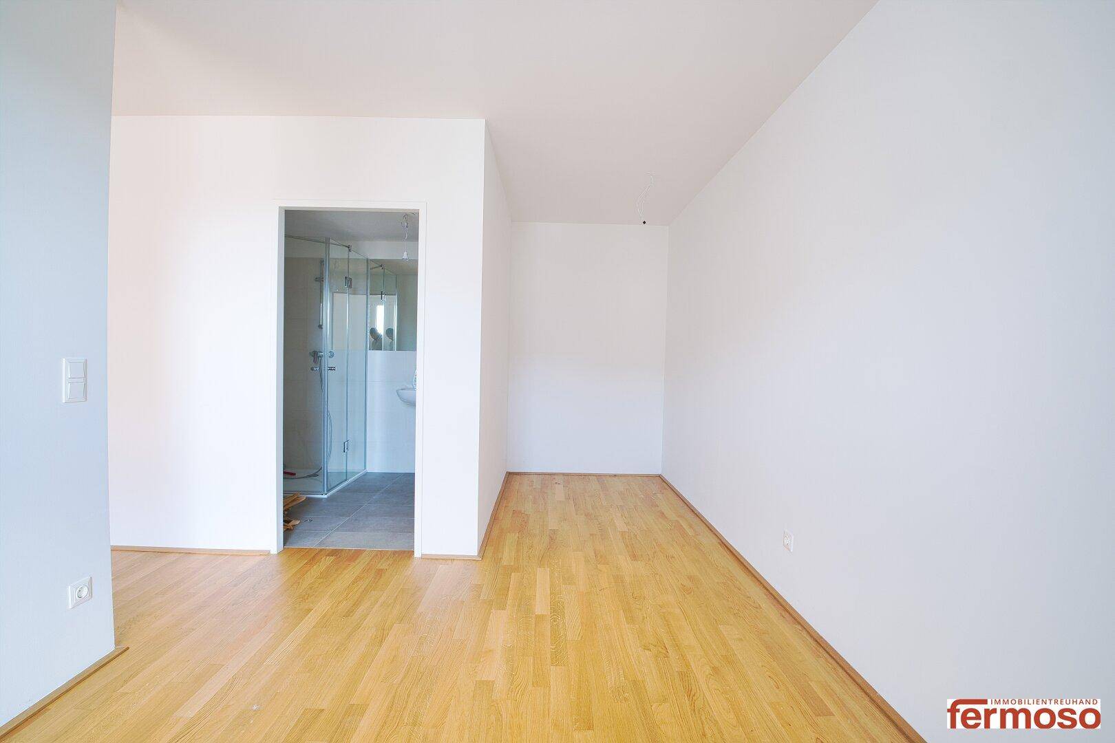Leerer Raum mit Parkettboden und Blick in ein modernes Badezimmer.