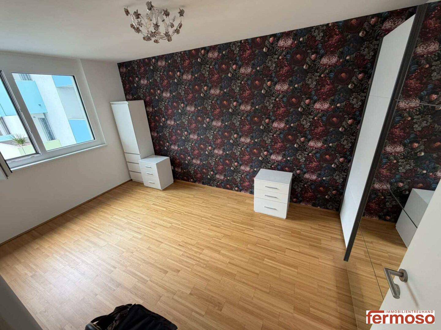 Schlafzimmer mit auffälliger floraler Tapetenwand, Holzboden und weißer Einrichtung.
