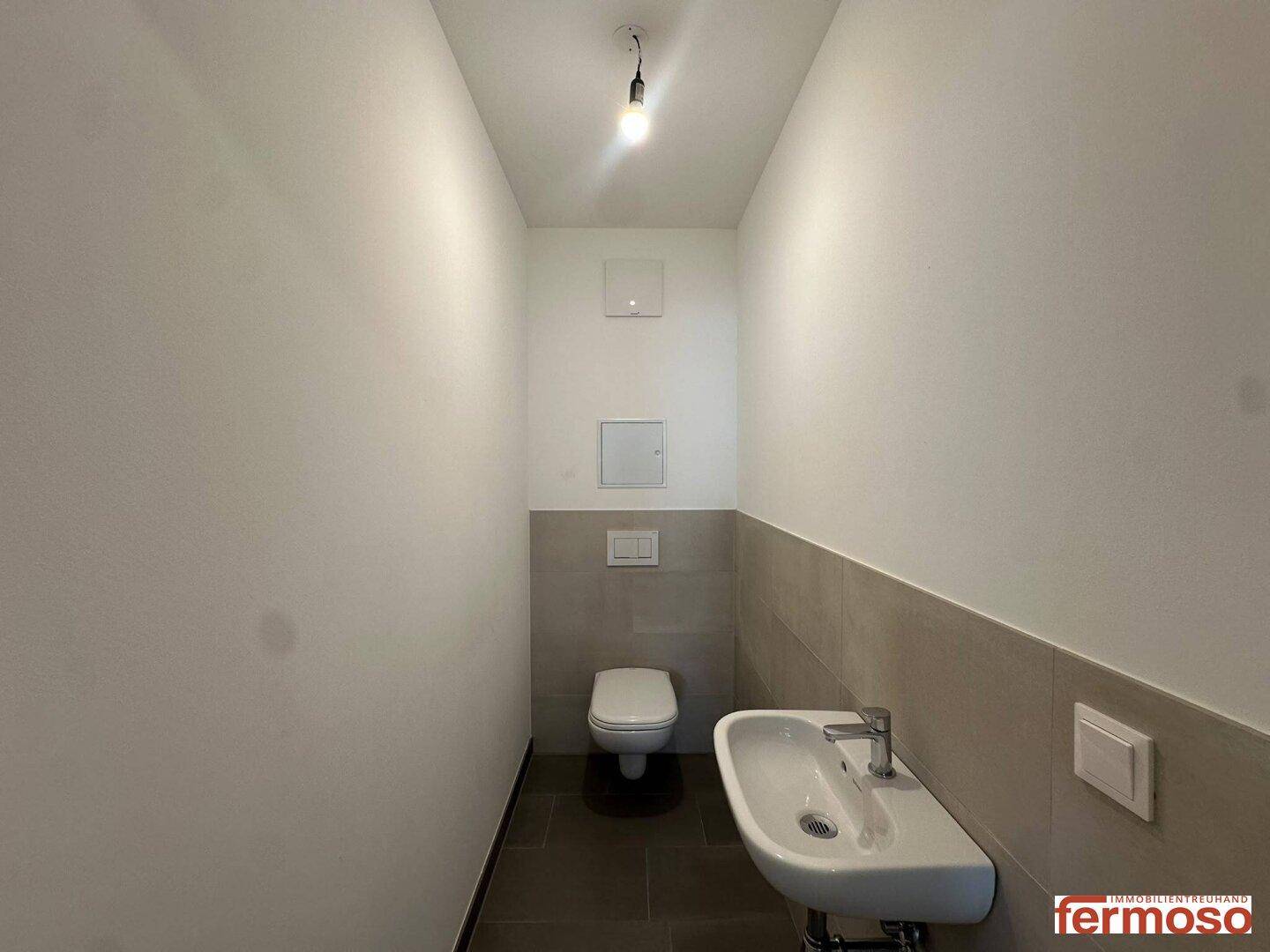 Modernes Gäste-WC mit Wand-WC, kleinem Waschbecken und hellen Fliesen an Wänden und Boden.