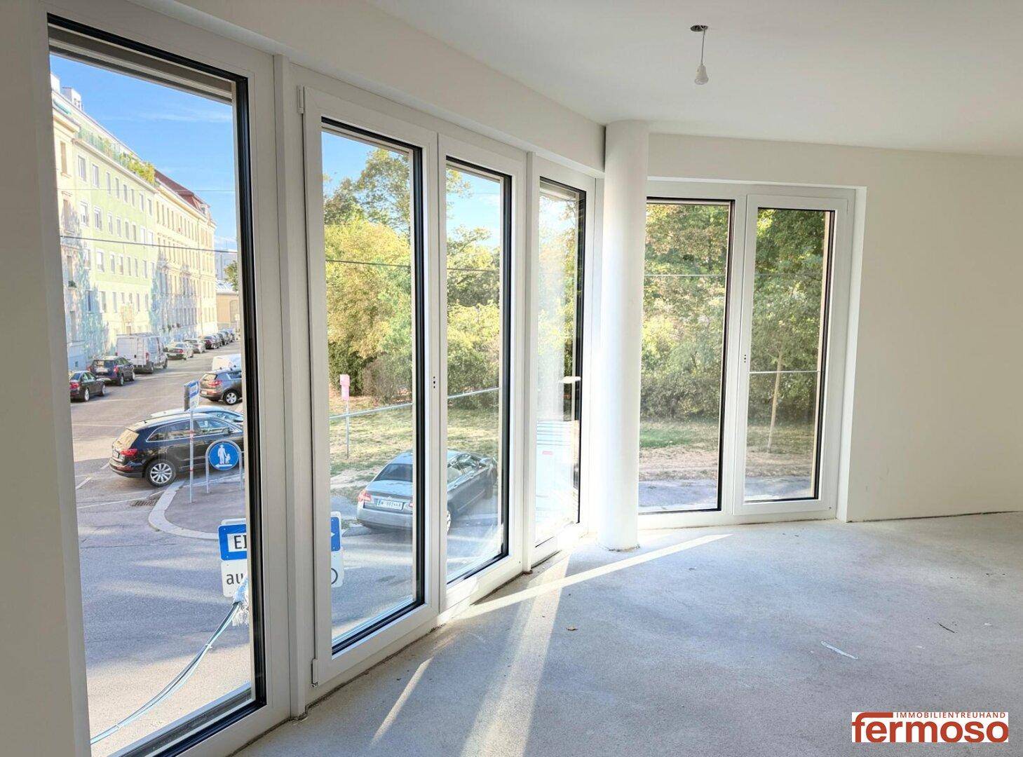 Heller Raum mit bodentiefen Fenstern, die einen Blick auf die Straße und Bäume bieten.