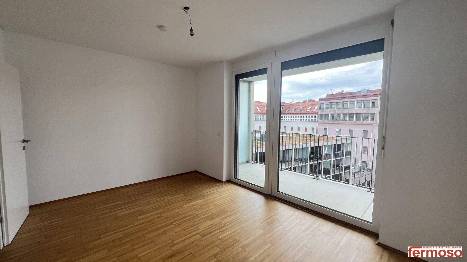 Heller Raum mit Holzboden und großen Fenstern, die Zugang zu einem Balkon bieten.