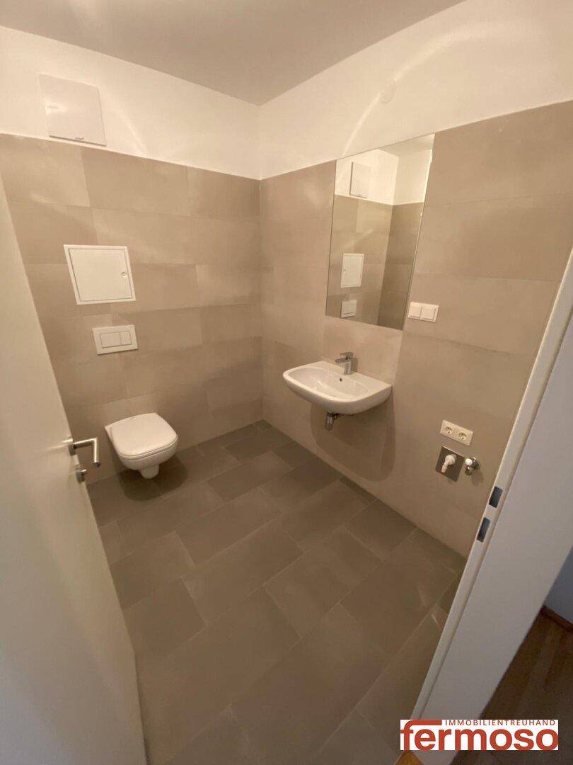 Modernes Gäste-WC mit Wand-WC, Waschbecken und großem Spiegel, hell gefliest.