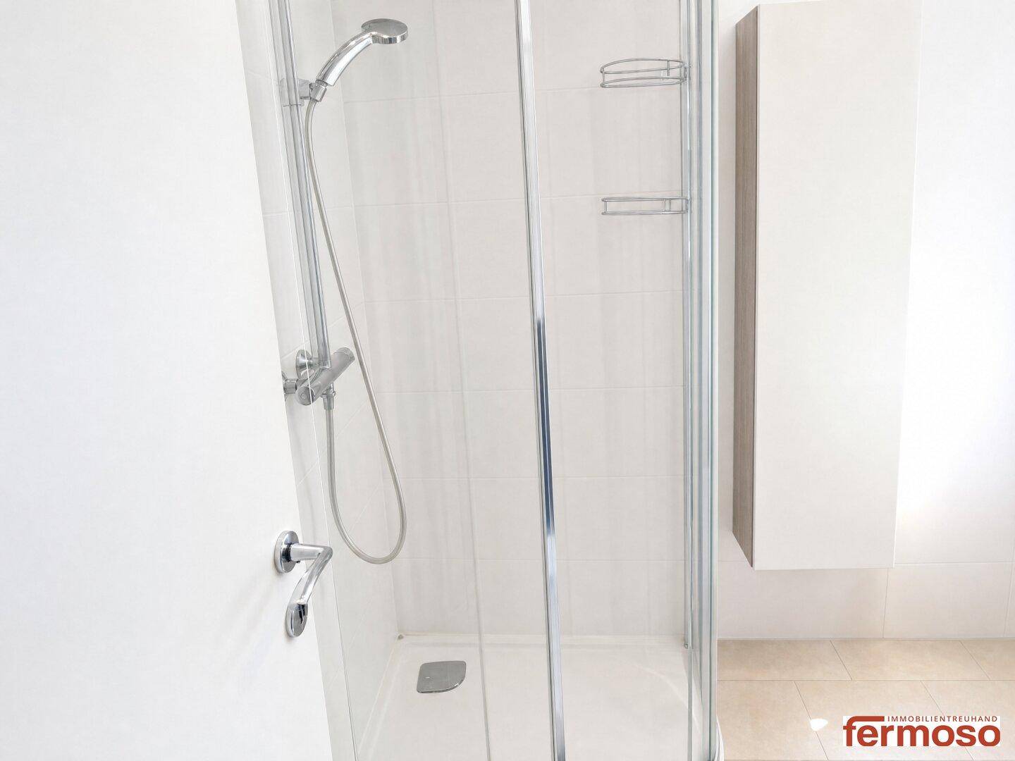 Moderne Duschkabine mit Glaswänden und Handbrause in einem hell gefliesten Badezimmer.