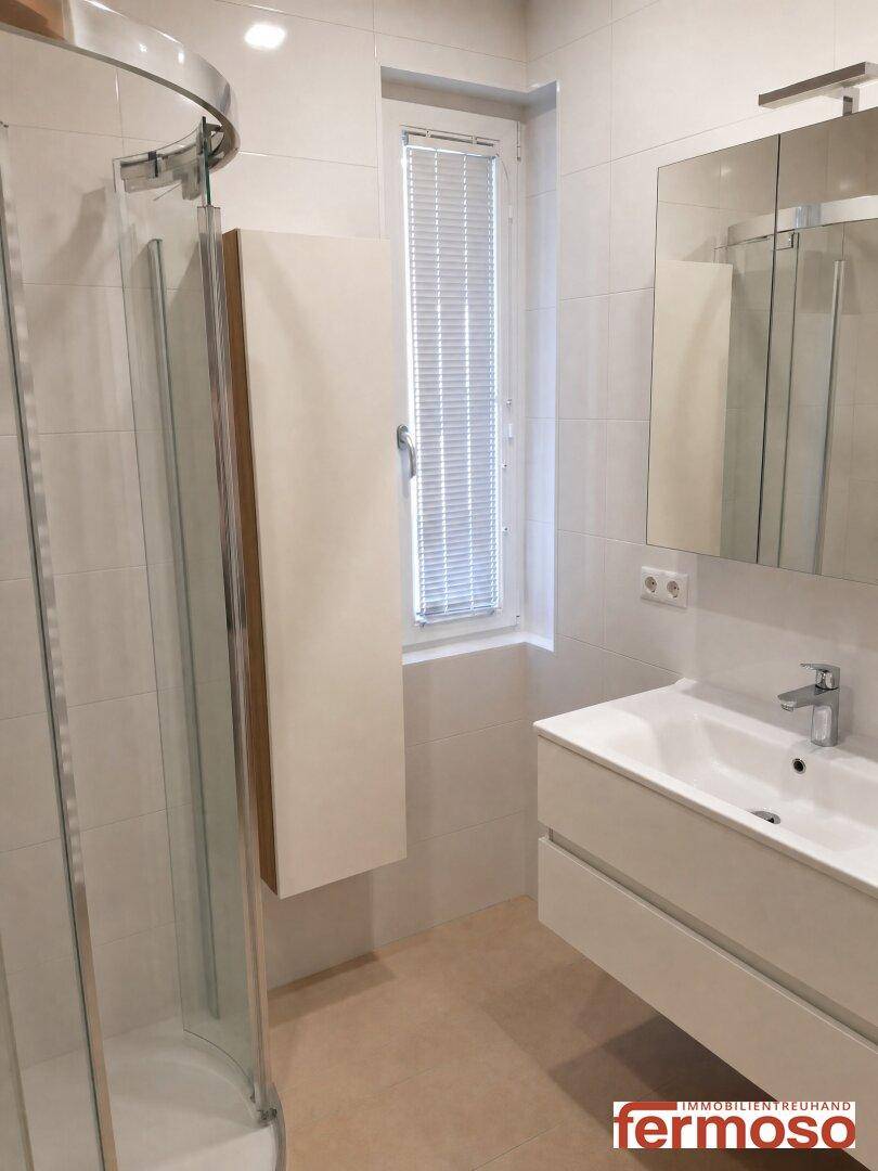 Helles Badezimmer mit Waschtisch, Spiegelschrank, Duschkabine und Fenster, modern gefliest.
