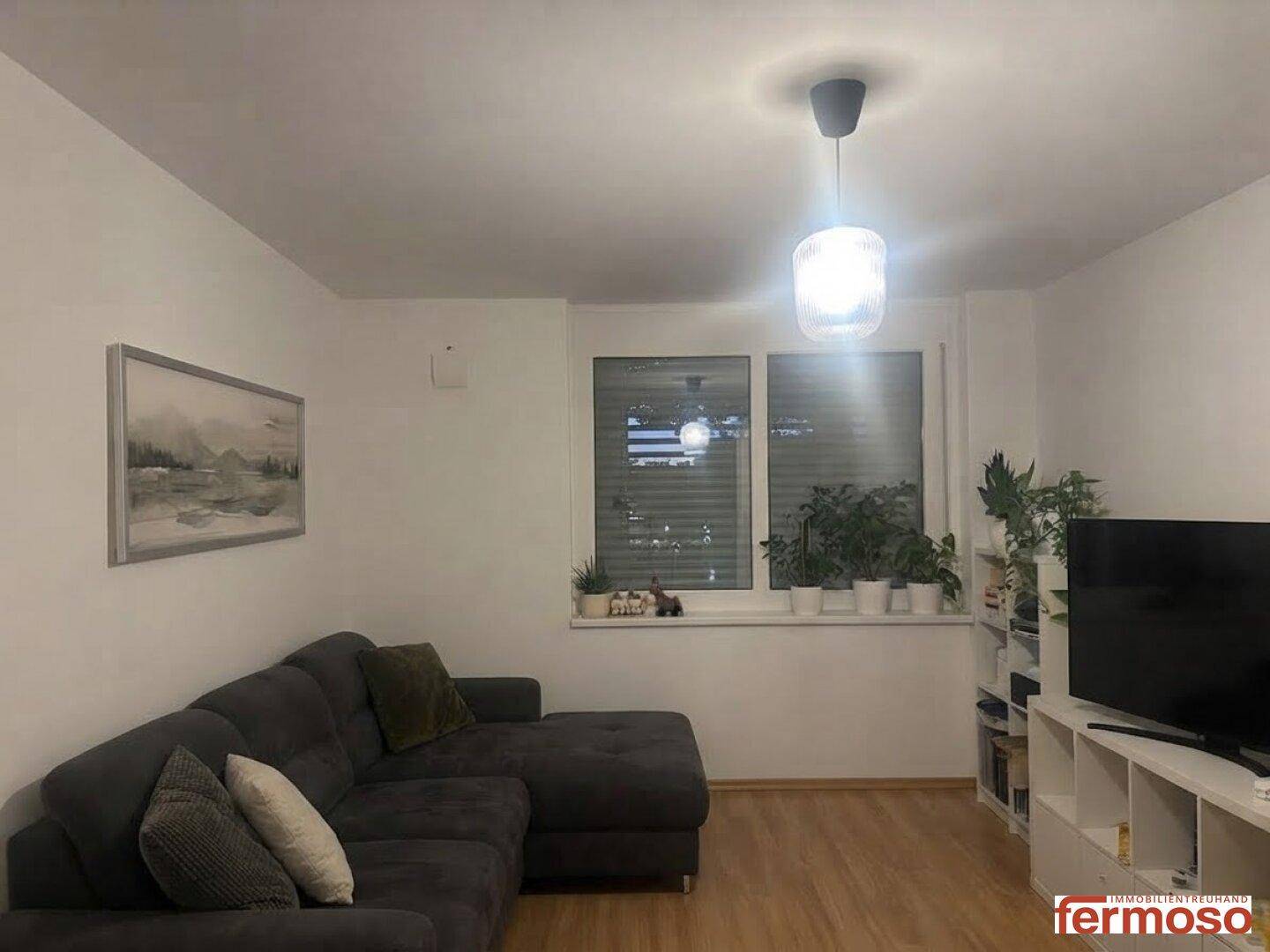 Gemütliches Wohnzimmer mit dunklem Ecksofa, Holzboden und Blick auf Pflanzen am Fenster.