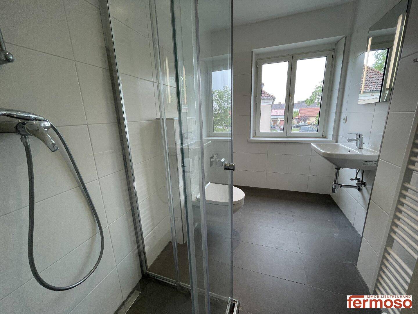 Helles Badezimmer mit Fenster, Duschkabine, Waschbecken und modernen grauen Fliesen.