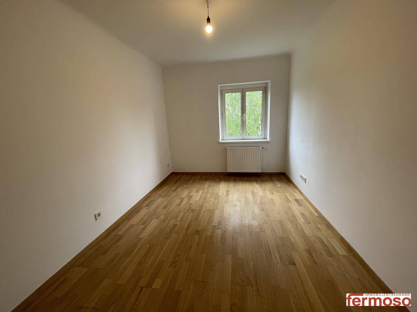 Helles Zimmer mit Parkettboden und einem Fenster, das einen Blick ins Grüne ermöglicht.