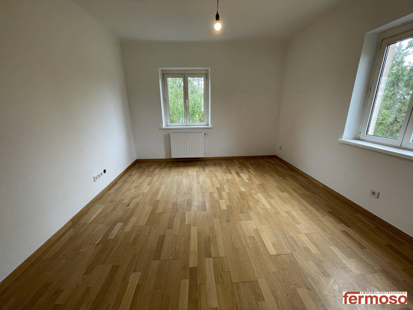 Geräumiges Schlafzimmer mit zwei Fenstern, die eine grüne Aussicht bieten, und hellem Parkettboden.
