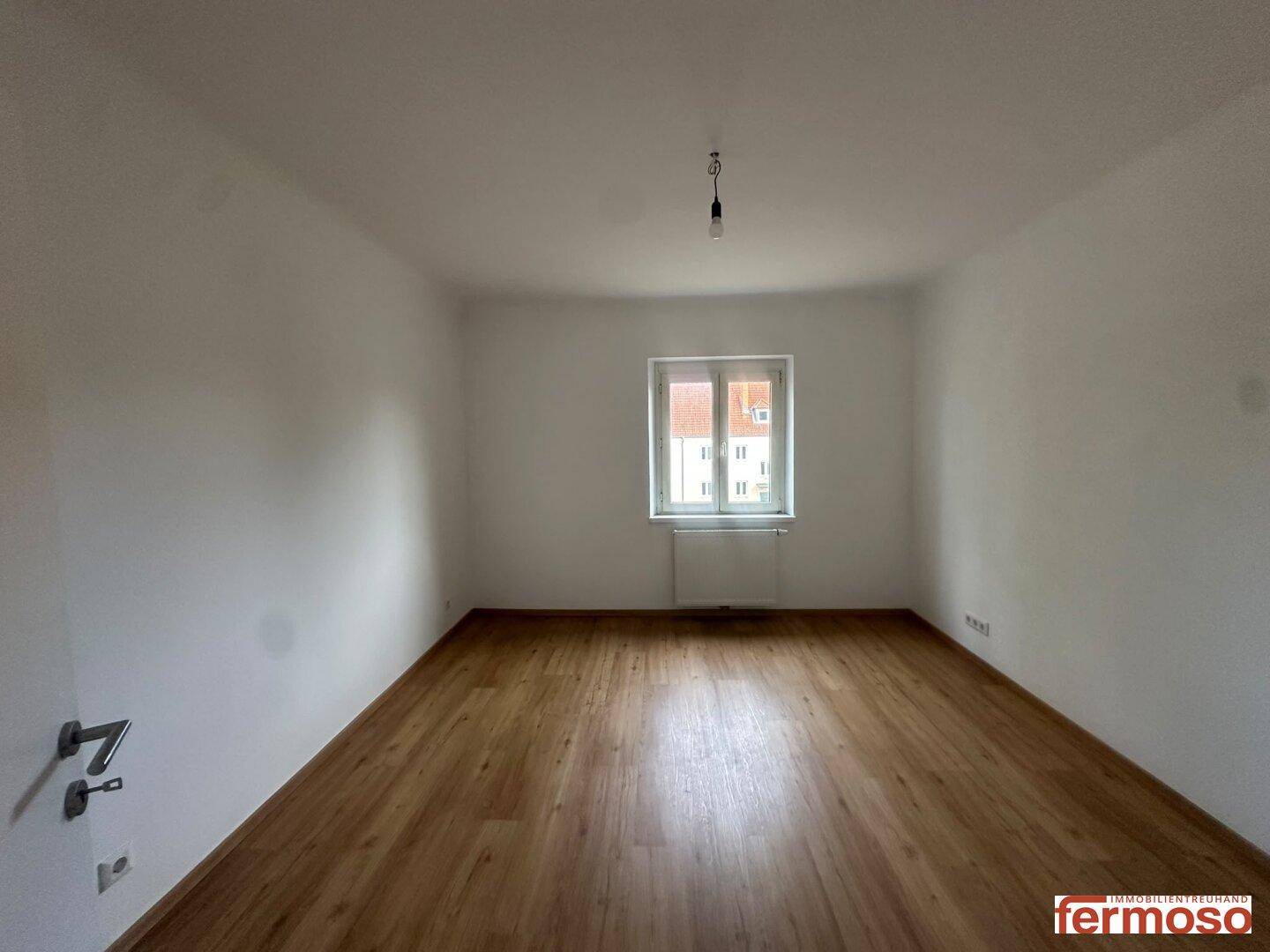 Leeres Zimmer mit hellem Laminatboden und weiß gestrichenen Wänden, bereit zur individuellen Gestaltung.