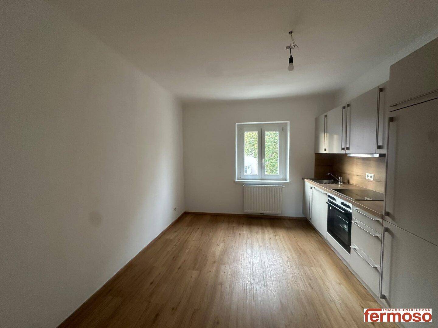 Offener Wohnbereich mit integrierter Küche, hellem Laminatboden und großem Fenster.