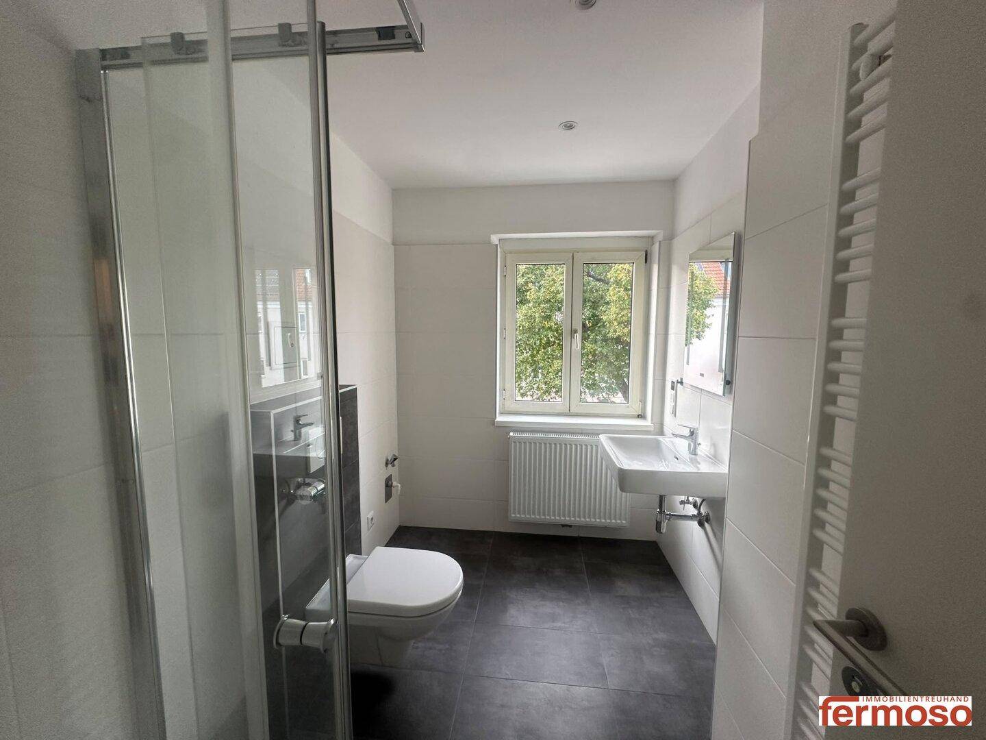 Modernes Badezimmer mit Dusche, WC, Waschbecken und Fenster, ausgestattet mit dunklen Bodenfliesen.