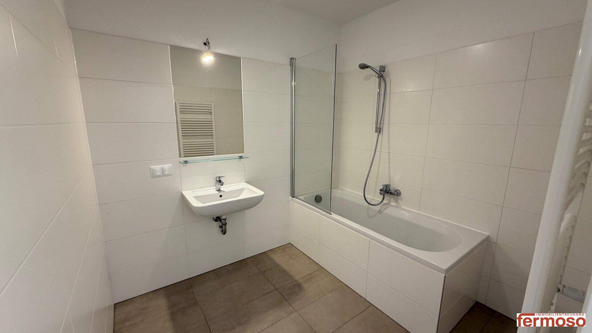Helles Badezimmer mit weißen Fliesen, Badewanne mit Duschwand und einem Waschbecken.