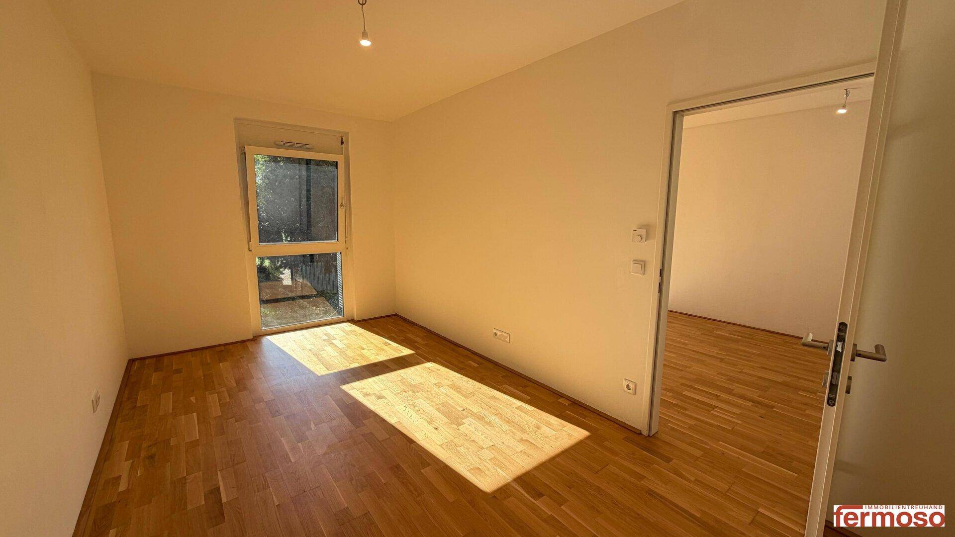 Leeres Zimmer mit hellem Parkettboden, großem Fenster und viel natürlichem Sonnenlicht.